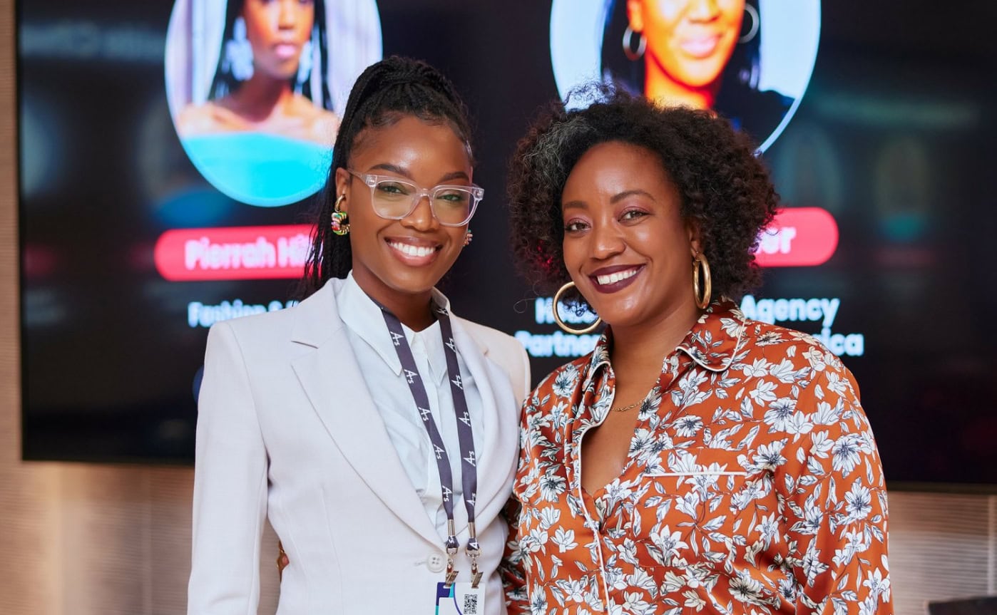 Two women smiling warmly stand together in front of a digital screen displaying their images and names. One wears a white suit; the other, a floral blouse.
