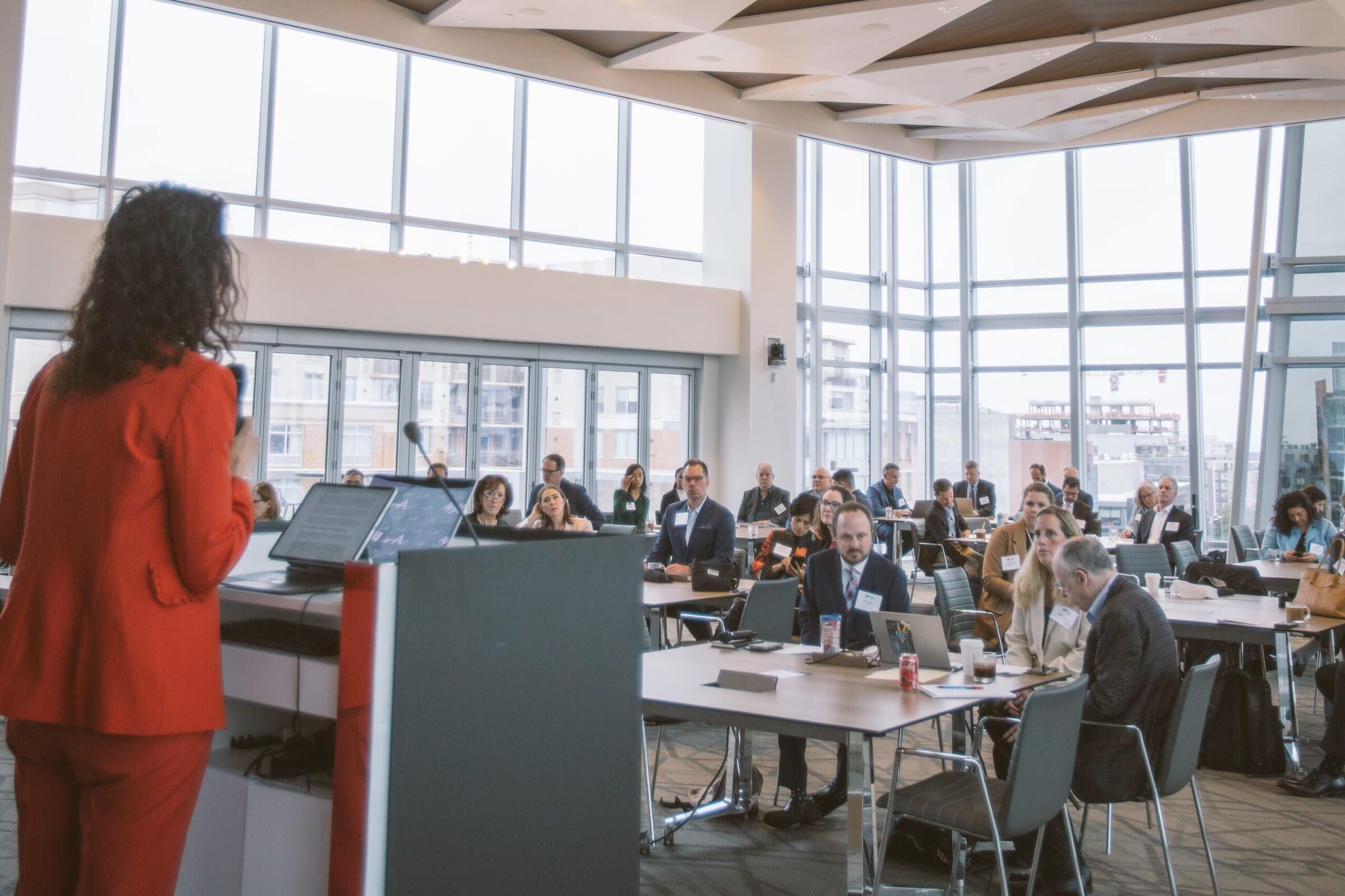 A person in a red suit speaks at a podium with a microphone, facing an audience seated at round tables in a bright, modern conference room with large windows.