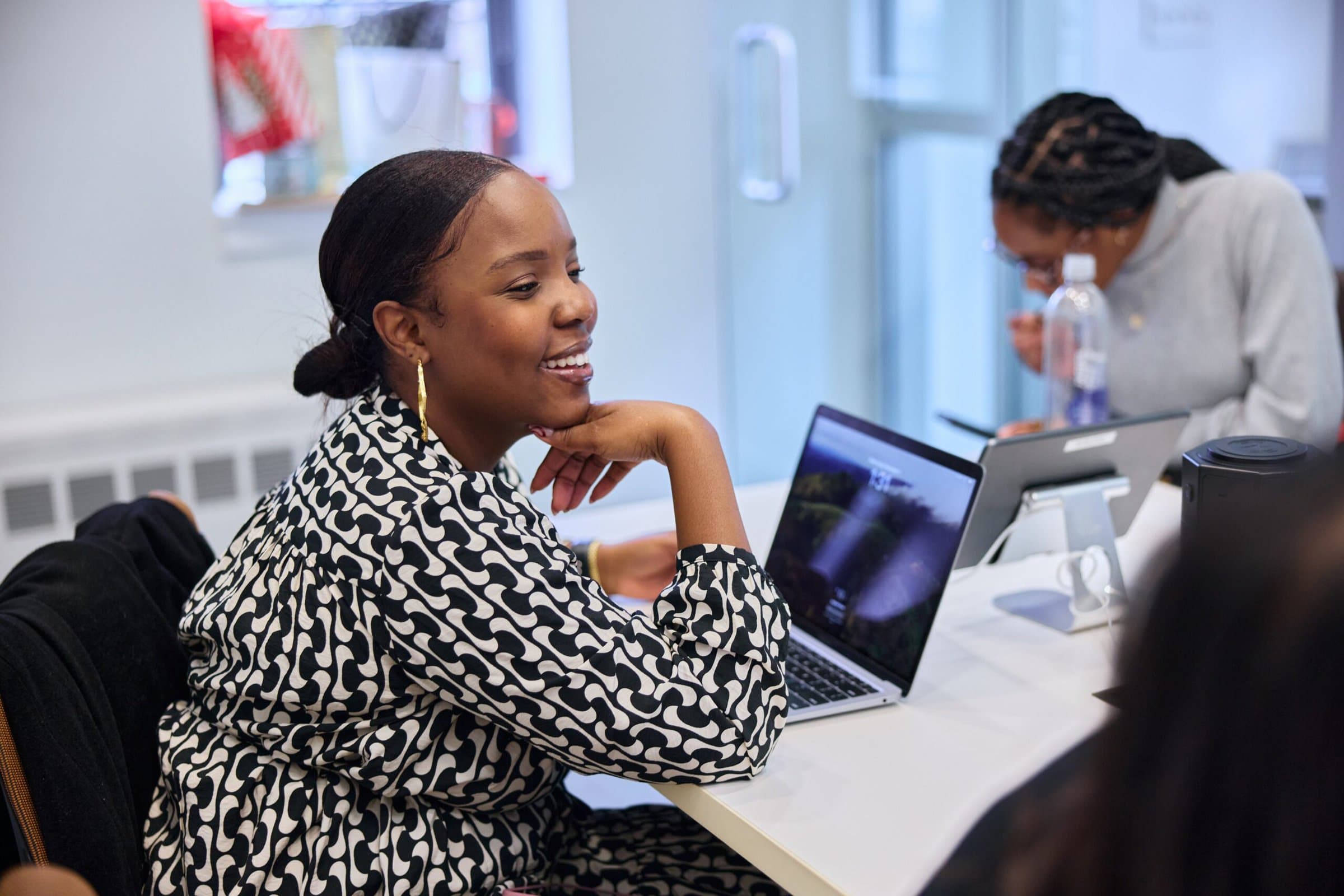 A cheerful woman in a bold black-and-white patterned outfit is seated at a table, resting her chin on her hand while looking to the side. Behind her, another person is busy with a tablet, adding a collaborative atmosphere.