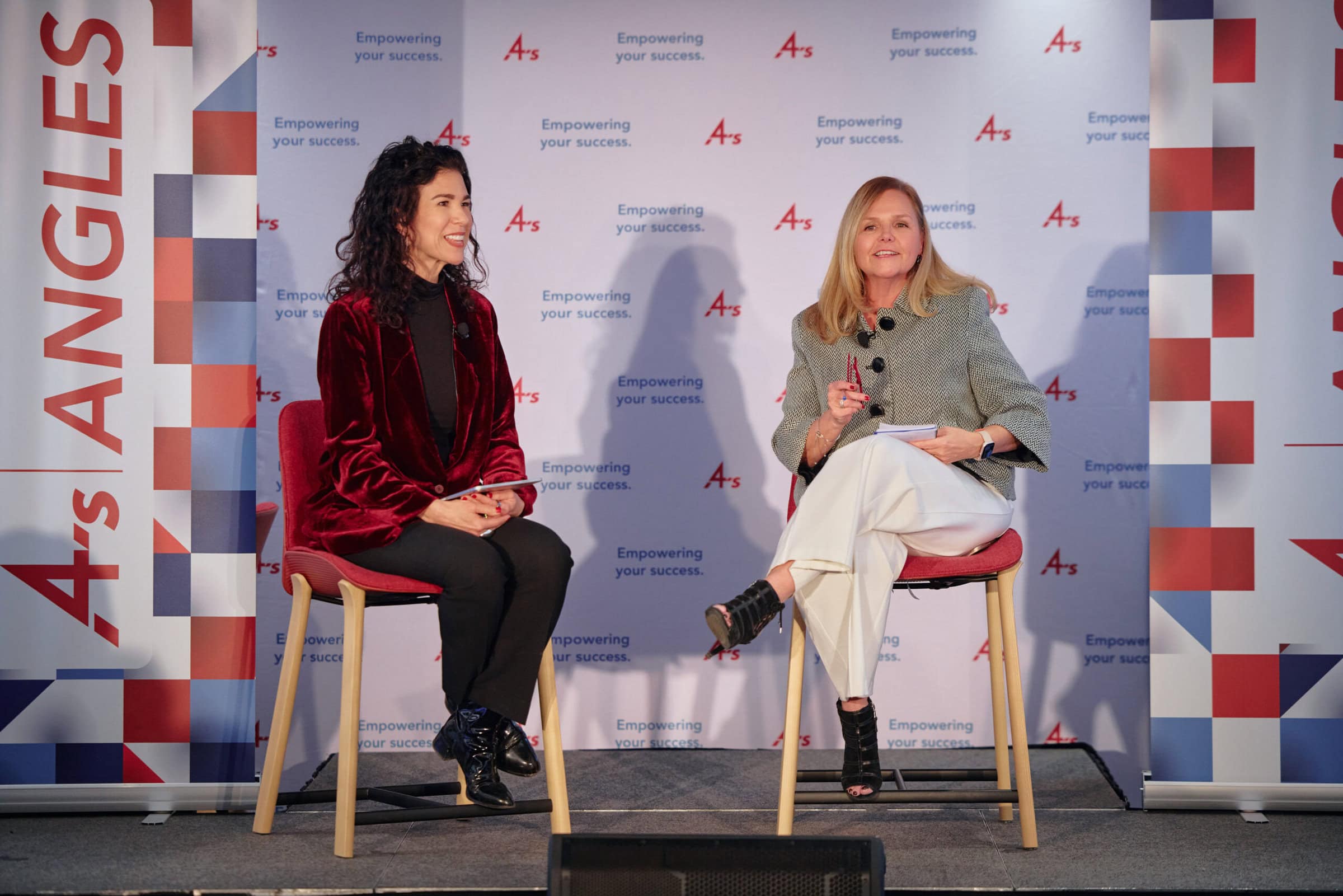 Two women sit on stage at a conference, smiling and engaged in conversation. Banners display the 4A's logo and the phrase "Empowering your success."
