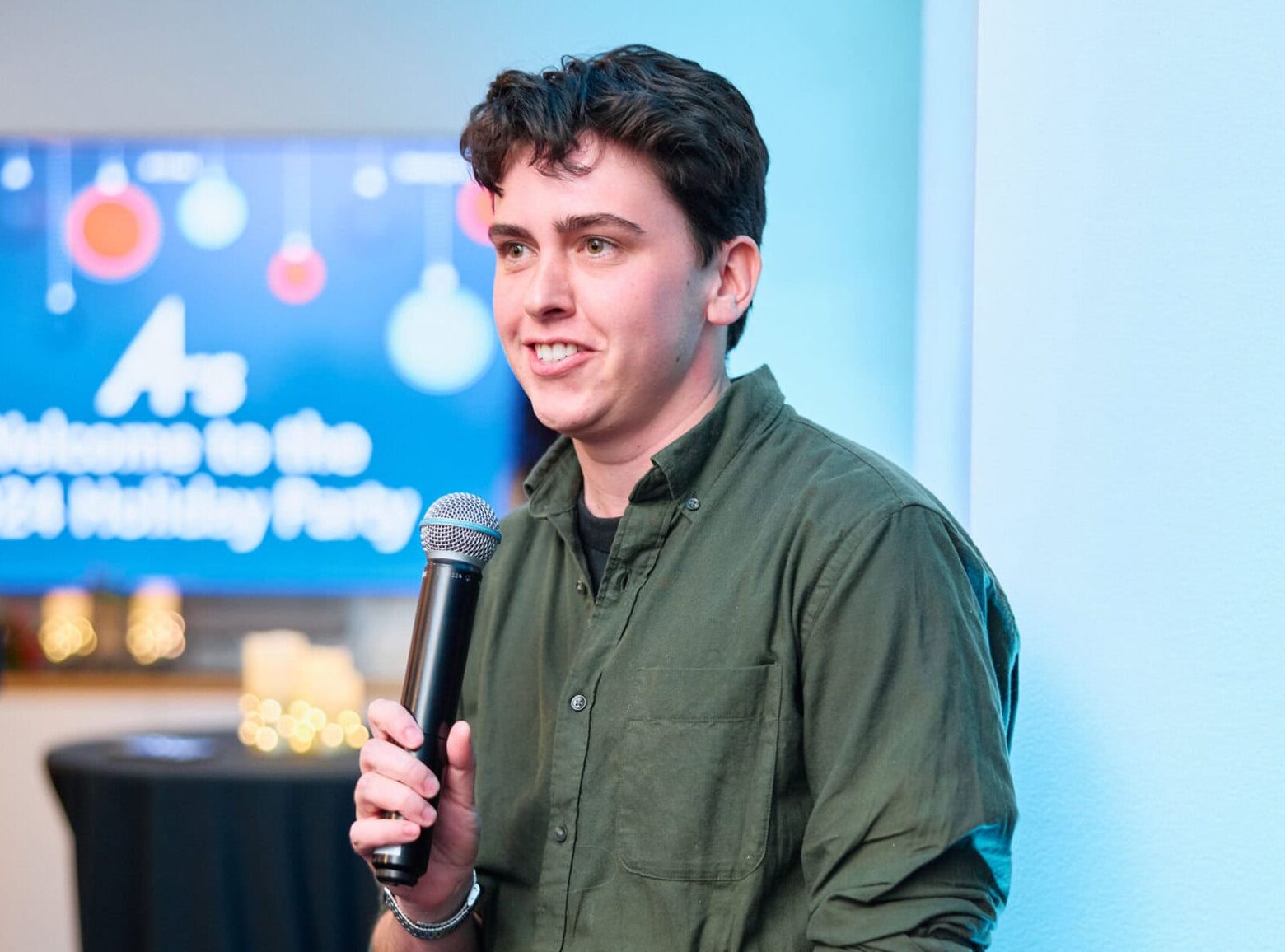 A young man in a green shirt speaks into a microphone at a festive event. A blurred sign in the background reads "Welcome to the 2024 Holiday Party."
