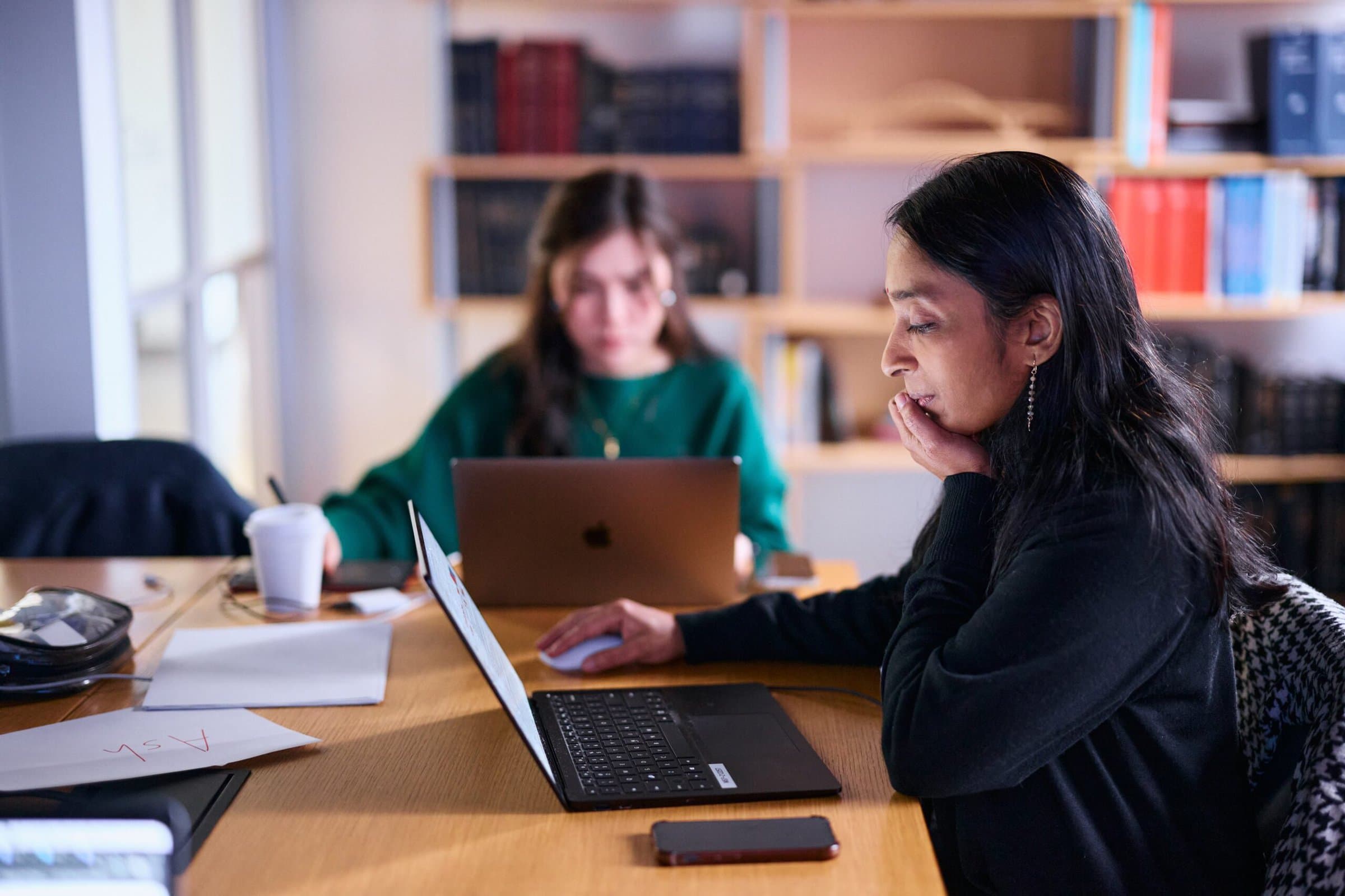 Two women focus intently on laptops in a library setting. One in the foreground is deeply engaged, while the other works in a blurred background. Cozy atmosphere.
