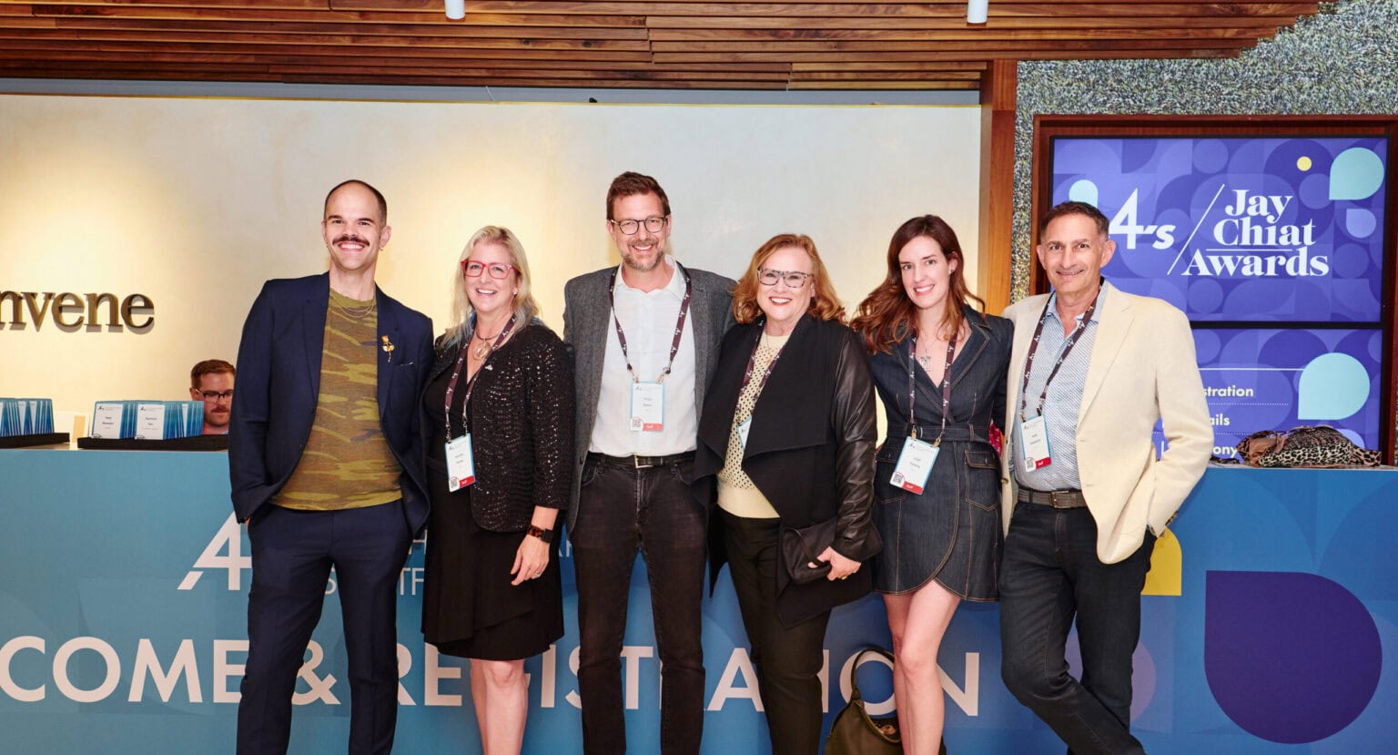Evoto A group of six people smiling and posing at an event. They stand in front of a reception desk and a sign that reads "4As Jay Chiat Awards." The mood is lively and professional.