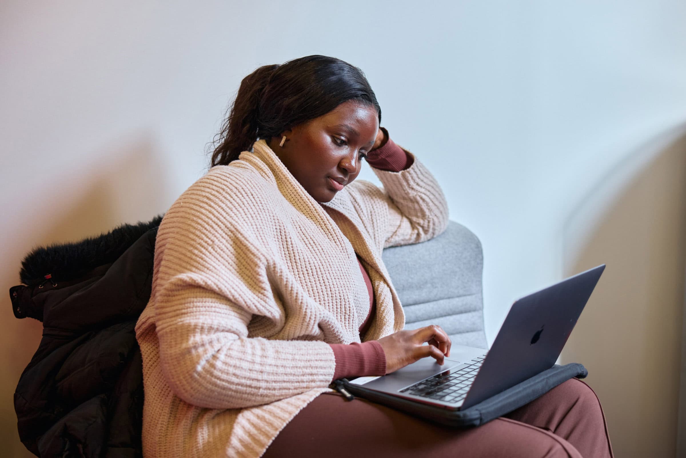 A woman in a tan sweater and maroon pants sits on a gray couch, focused on typing on a laptop. Her expression is thoughtful and relaxed.