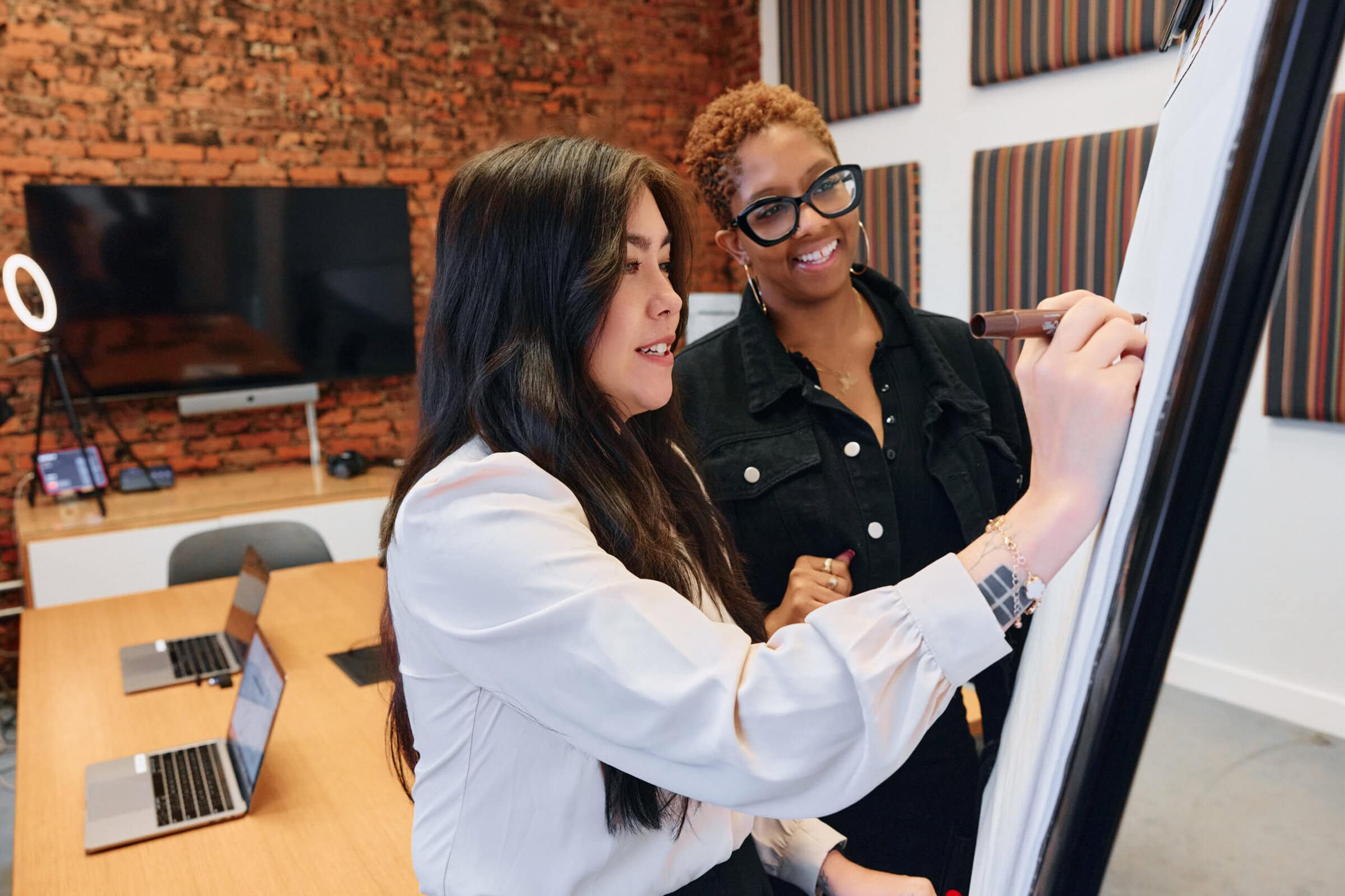 Two women collaborate enthusiastically at a flipchart in a modern office. Laptops and a large screen are visible, creating a productive atmosphere.